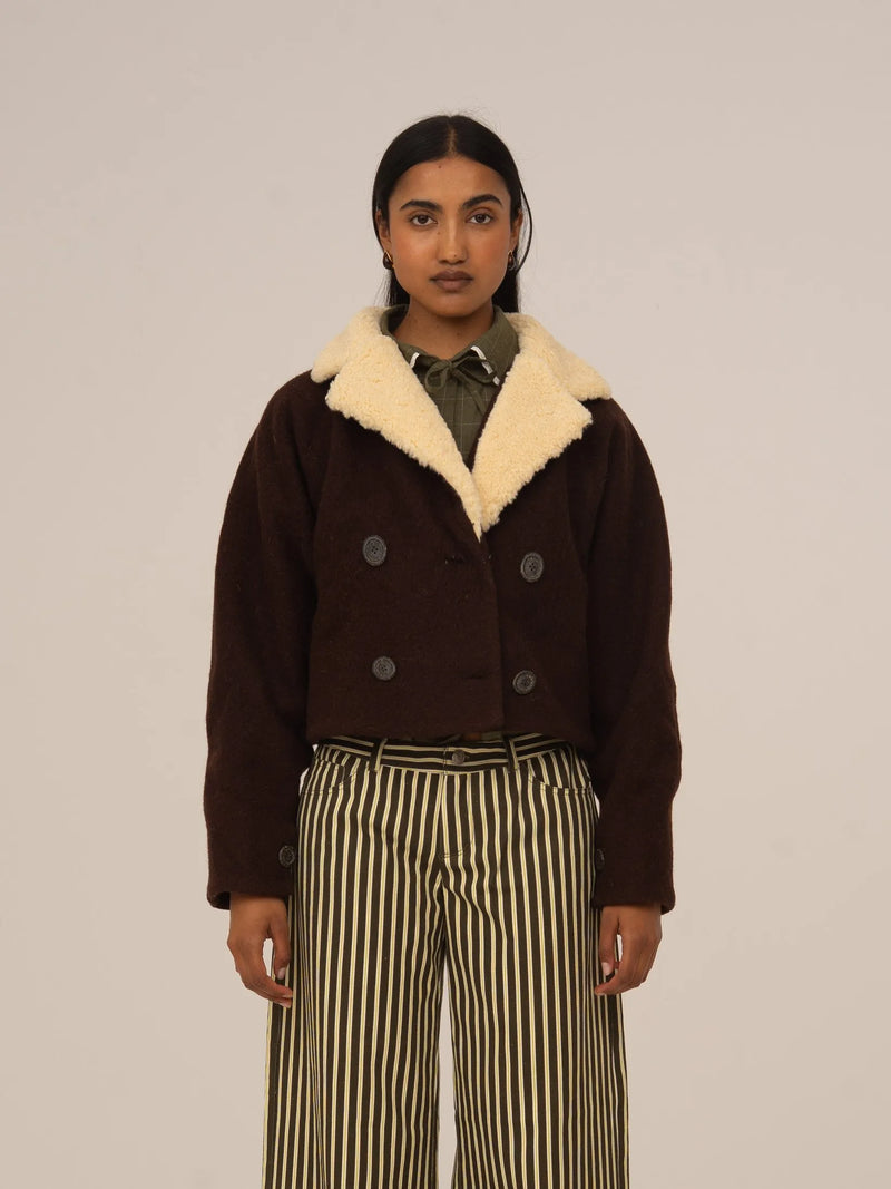 Chaqueta corta marrón de corte cropped con cuello de borreguito beige y doble botonadura, presentada sobre modelo en estudio con fondo neutro.