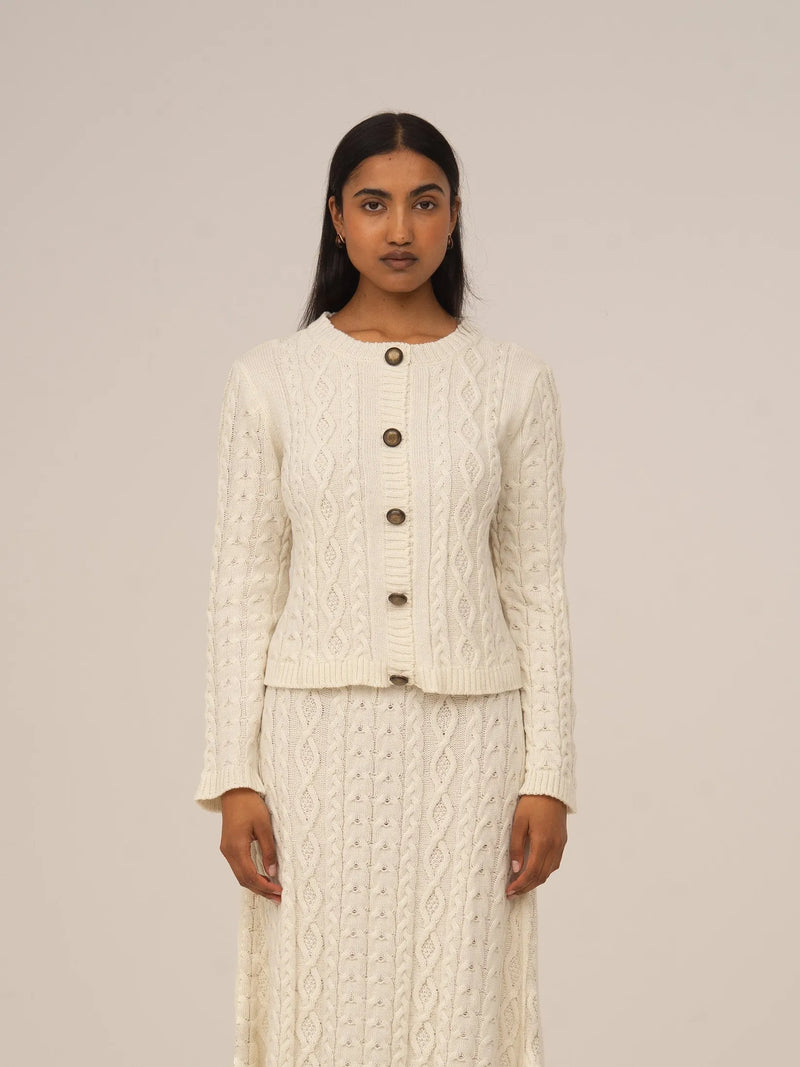 Cárdigan blanco de punto con botones frontales y textura trenzada, presentado sobre modelo en estudio con fondo neutro.