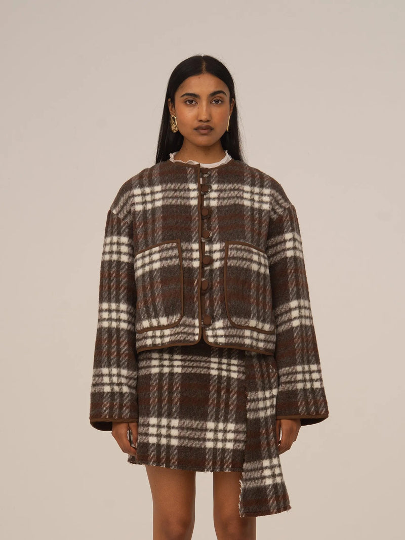 Chaqueta corta de cuadros marrón con botones frontales y cuello redondo, combinada con falda a juego, presentada sobre modelo en estudio con fondo neutro.