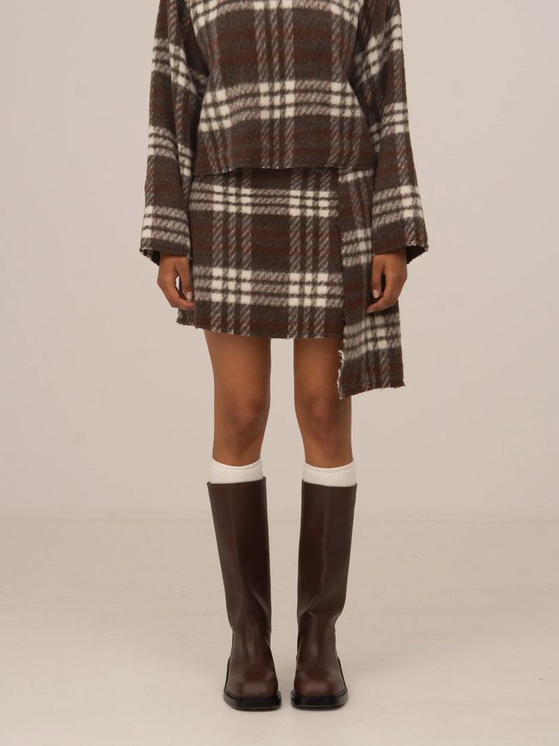 Minifalda marrón de cuadros con patrón tartán, combinada con botas altas, presentada sobre modelo en estudio con fondo neutro.