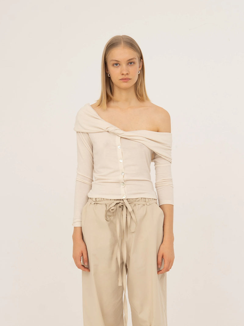 Top blanco de punto con diseño off-shoulder, detalle drapeado cruzado en el escote y cierre frontal con botones. Silueta ajustada y manga larga, confeccionado en jersey suave para un ajuste cómodo y femenino.