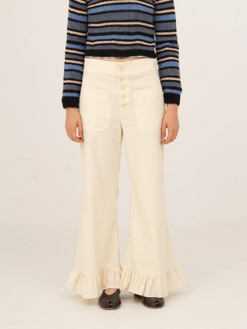 Pantalón blanco de pana con corte recto y pernera ligeramente acampanada, cierre frontal con botones y bajo con volante, presentado sobre modelo en estudio con fondo neutro.