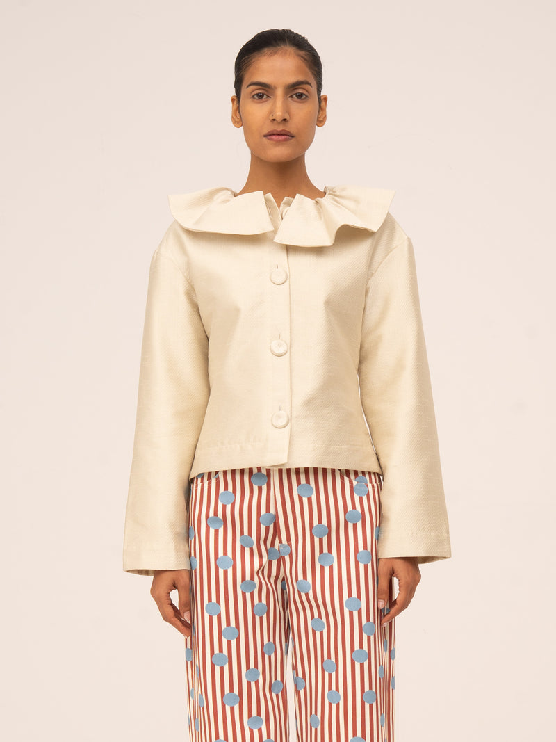 Chaqueta corta en color crema con ligero brillo, cuello amplio con volante y botones frontales, de estilo elegante.