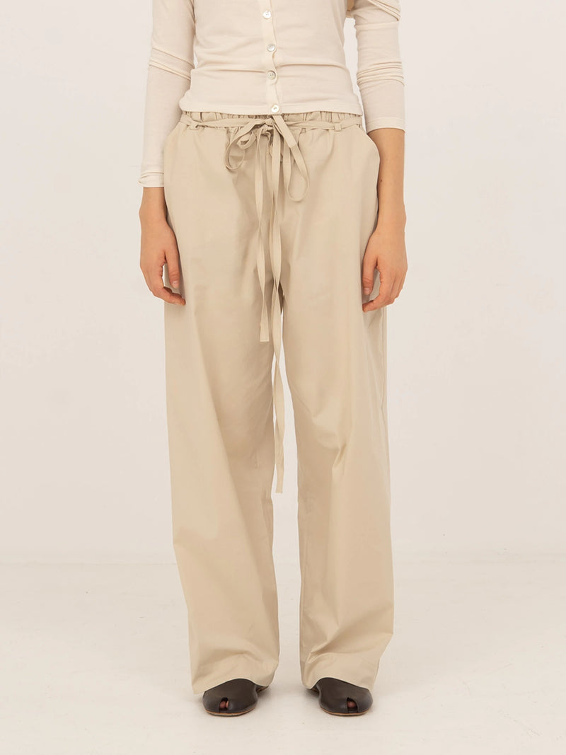 Pantalón beige de tejido fluido con corte recto y pernera larga, cintura elástica con lazada ajustable y caída suave, presentado sobre modelo en estudio con fondo neutro.