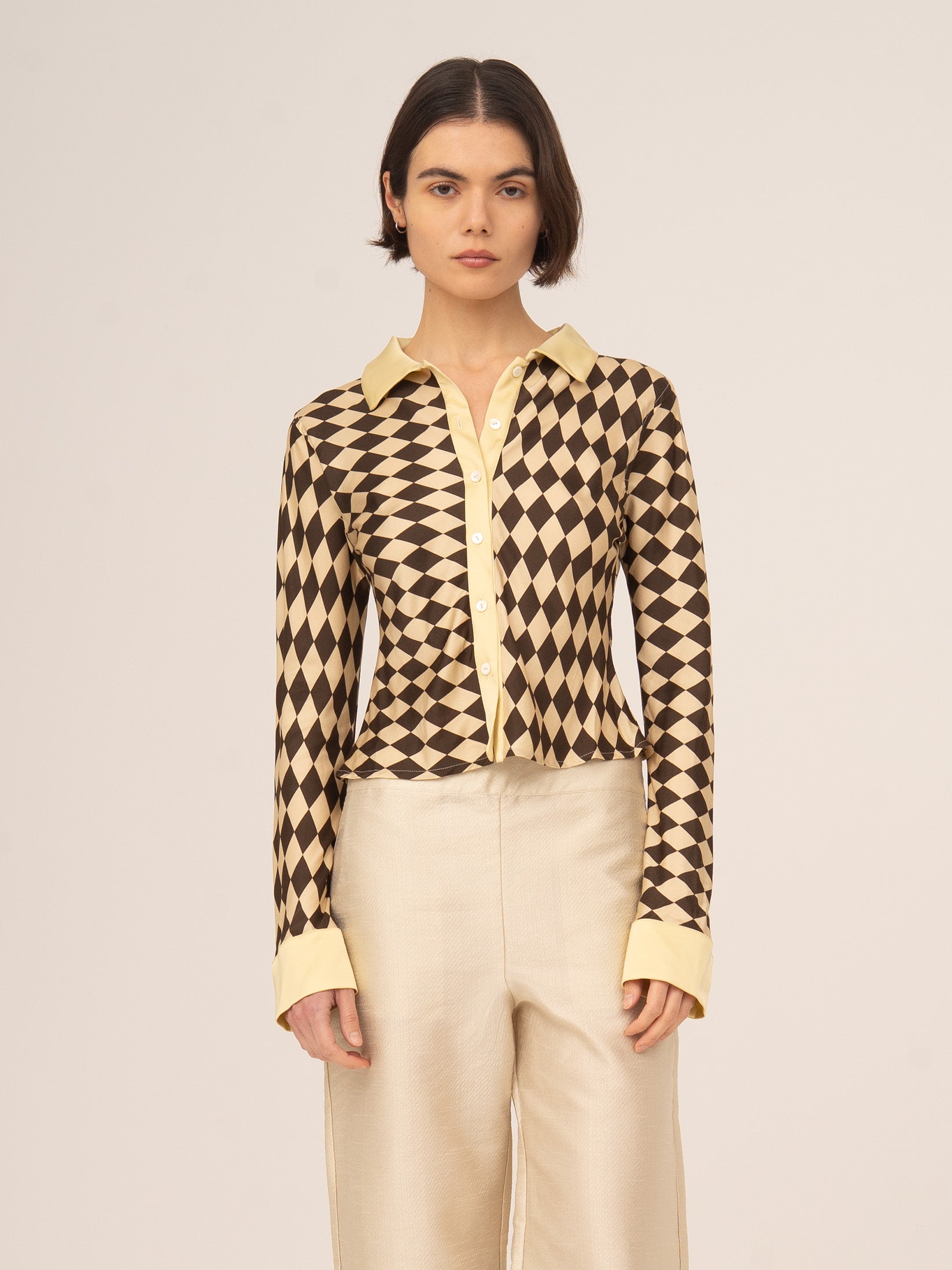 Modelo femenina vistiendo top de manga larga con estampado geométrico tipo argyle en tonos marrón y beige, combinado con pantalón claro, fotografiado de frente sobre fondo neutro.