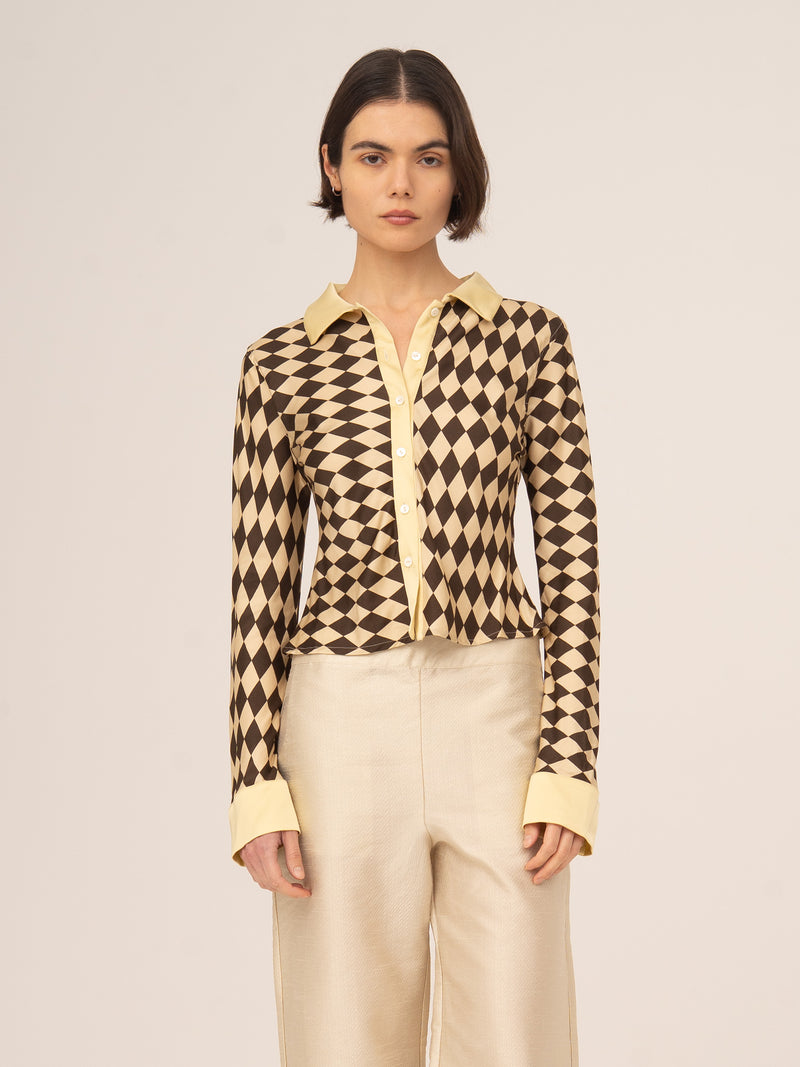 Modelo femenina vistiendo top de manga larga con estampado geométrico tipo argyle en tonos marrón y beige, combinado con pantalón claro, fotografiado de frente sobre fondo neutro.