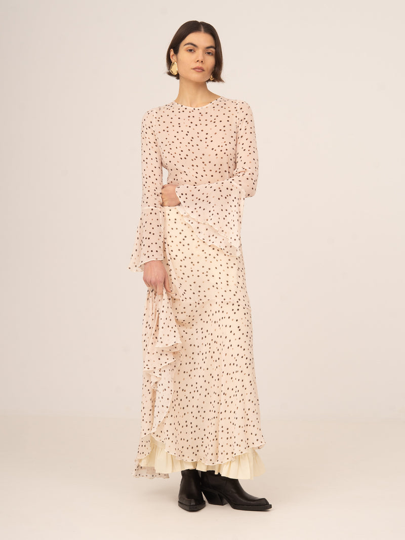 Modelo femenina de pie luciendo vestido largo fluido en tono melocotón con estampado de lunares, manga larga y corte relajado, con bajo ligeramente escalonado, combinado con botines negros sobre fondo claro y neutro.