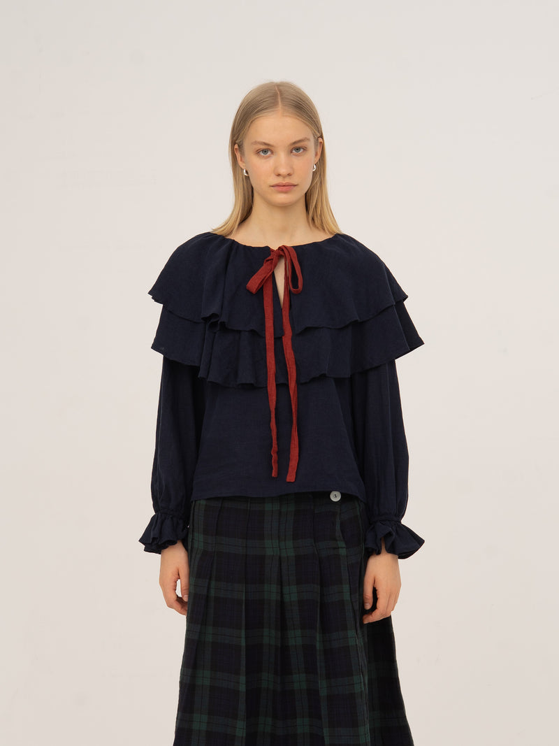 Blusa azul marino con capas de volantes en el pecho, lazada frontal en contraste rojo, manga larga con puño fruncido y caída fluida, presentada sobre modelo en estudio con fondo neutro.