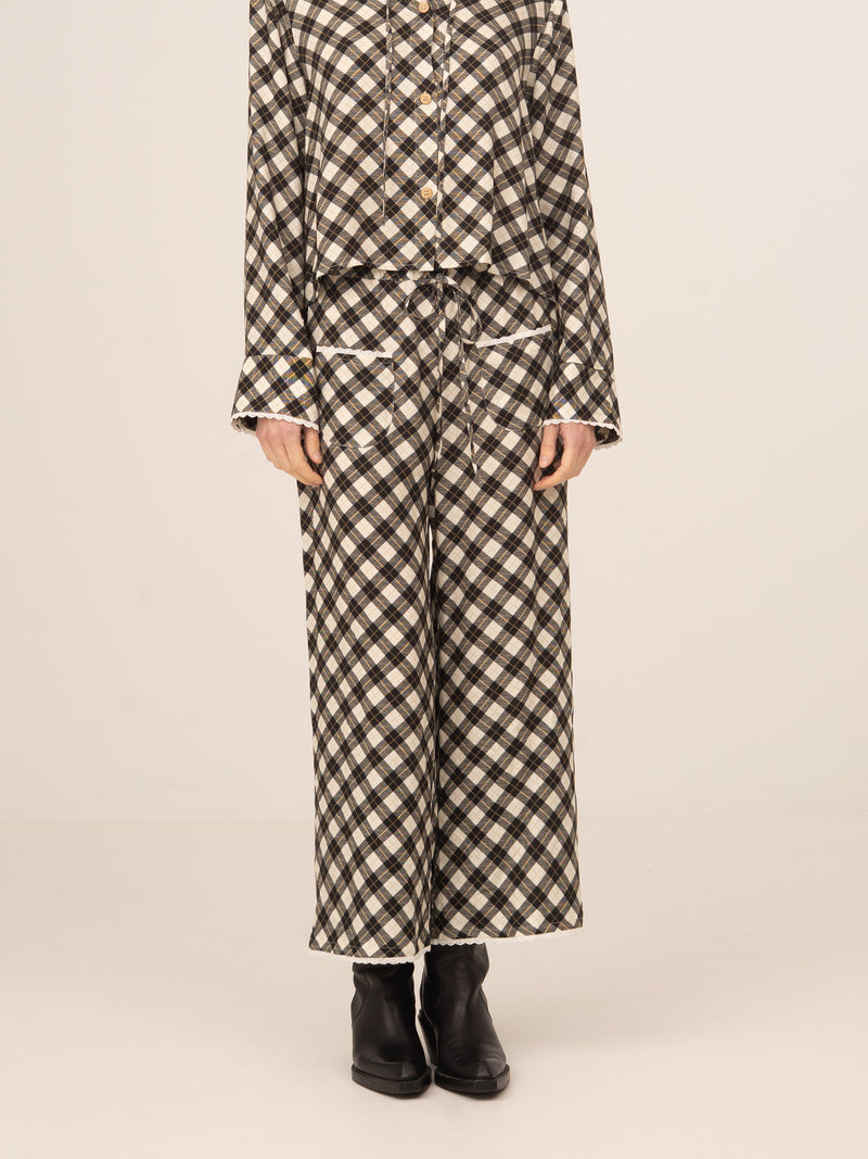 Modelo femenina de pie mostrando pantalón largo de corte recto y pernera amplia, con estampado geométrico tipo argyle en tonos neutros, cintura relajada y caída fluida, combinado con camisa a juego y botines negros sobre fondo claro y neutro.