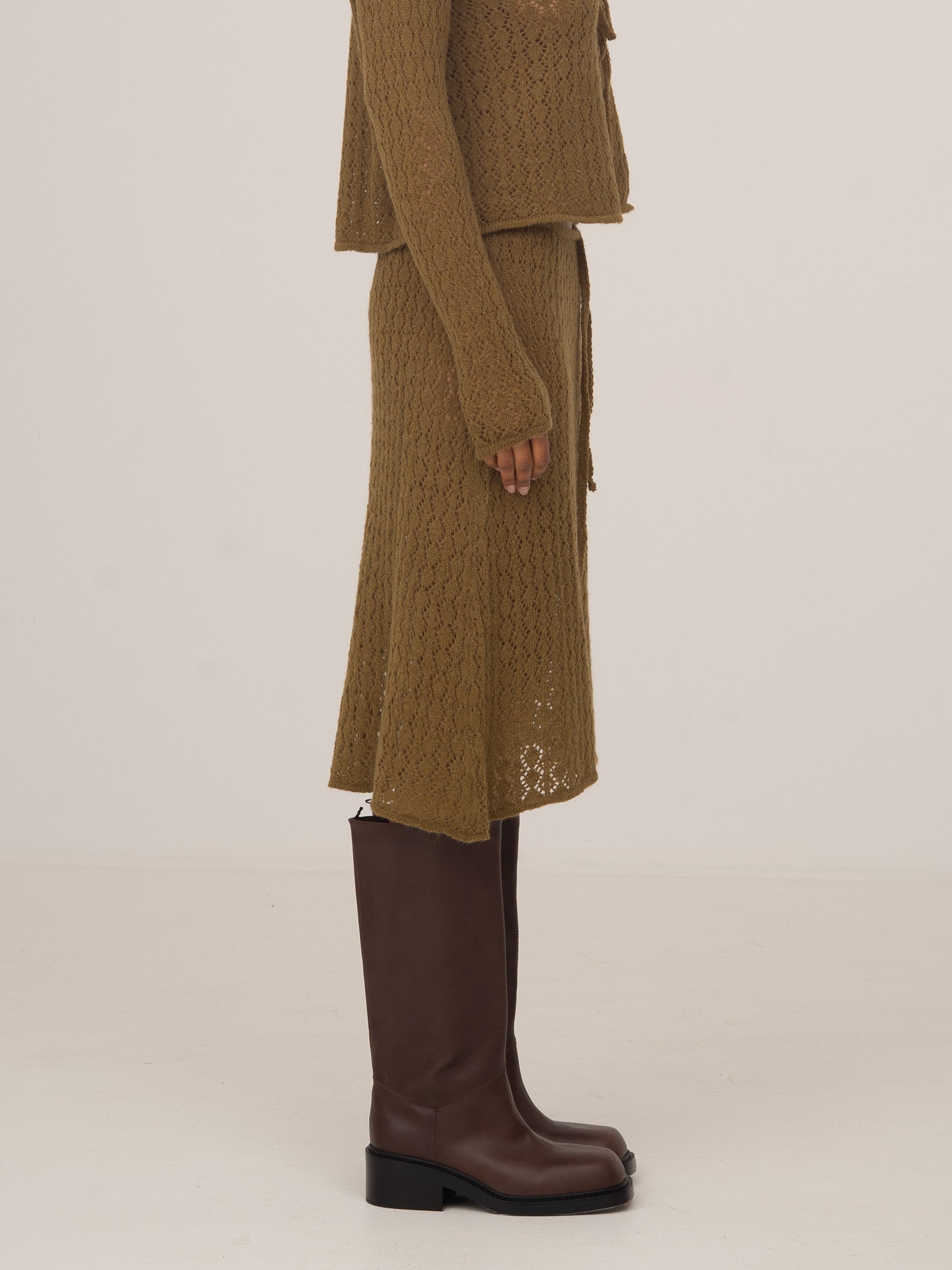 Vista lateral de falda midi de punto verde con textura trenzada, combinada con cárdigan a juego y botas altas marrones