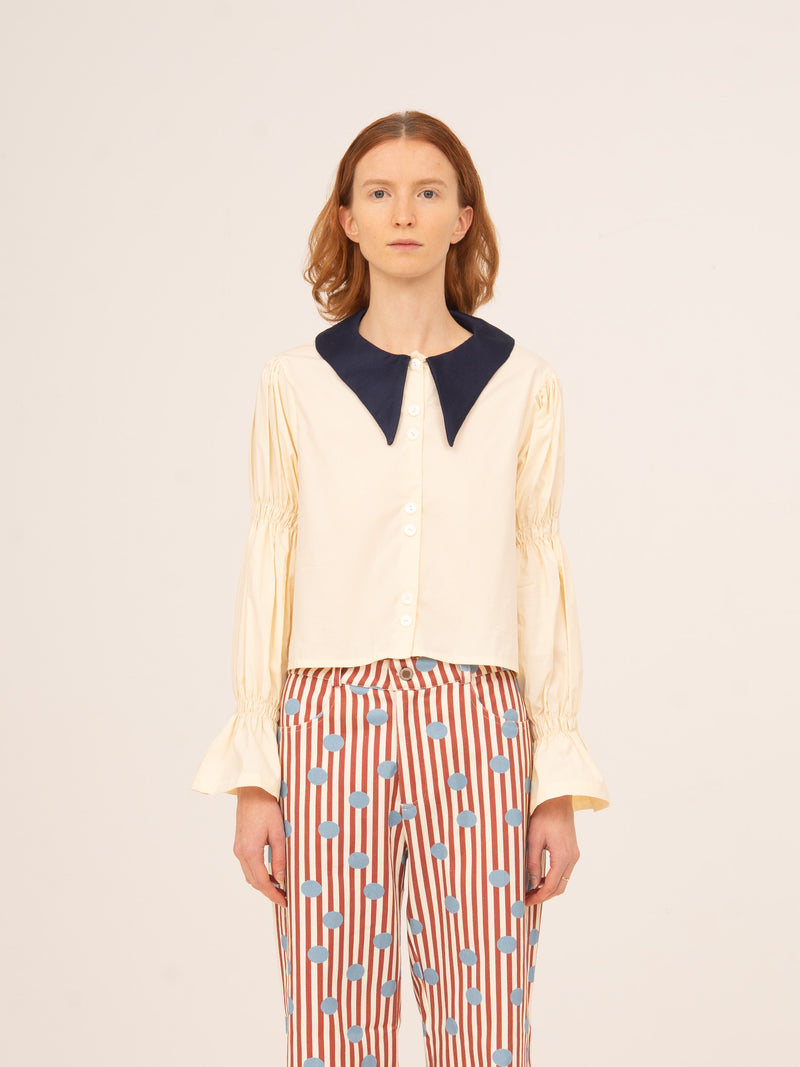 Blusa femenina en color blanco con cuello camisero en contraste azul marino y mangas largas con volumen y puños acampanados, diseño elegante y delicado ideal para looks sofisticados, conjuntos de invitada o estilismos especiales con un toque romántico.